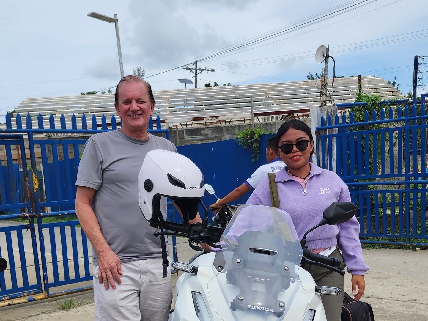 Traveller and a smiling woman with a white Honda ADV 160 by the blue airport fence