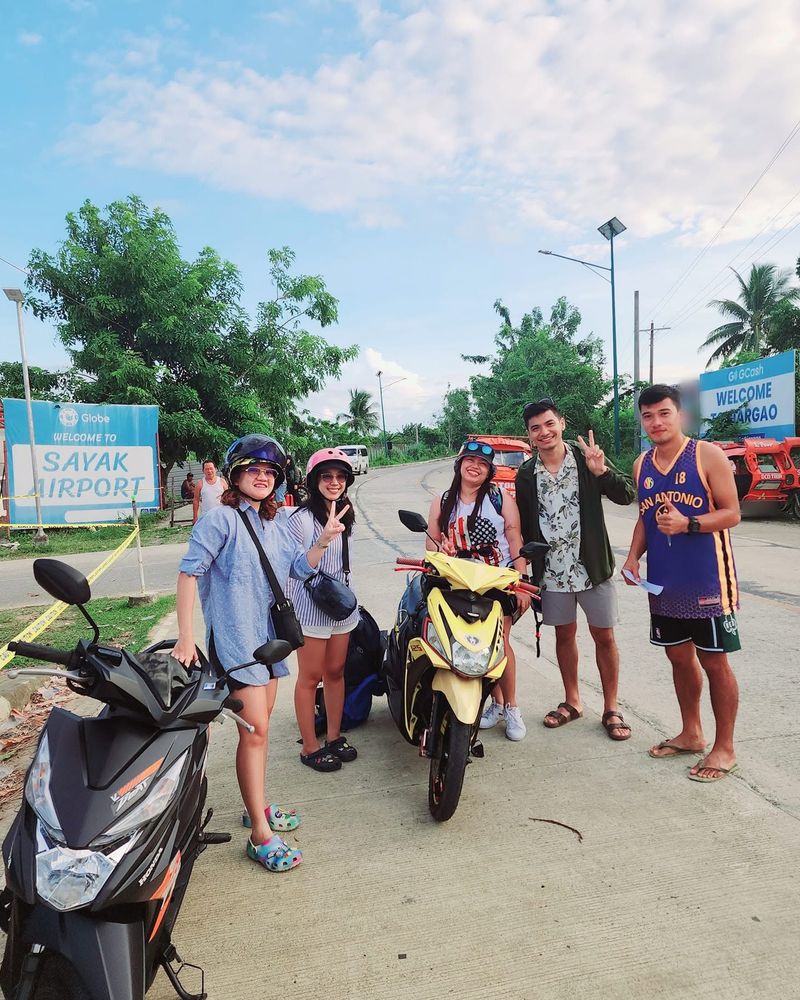 Smiling travellers with their rental scooters by the Welcome to Sayak Airport sign