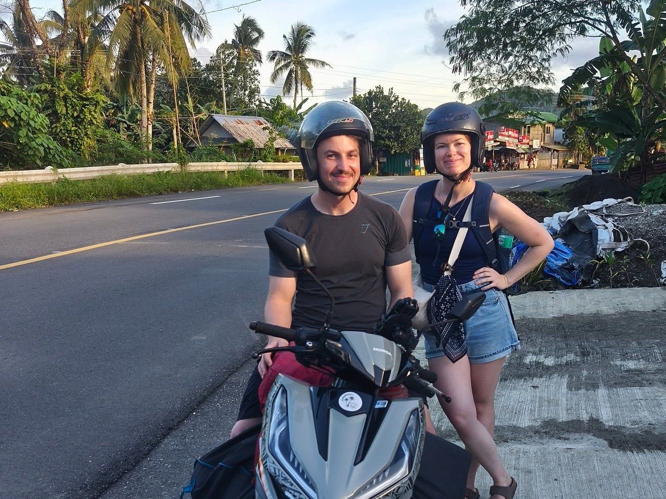 Smiling couple in helmets with a grey Honda Click on a highway in late-afternoon light