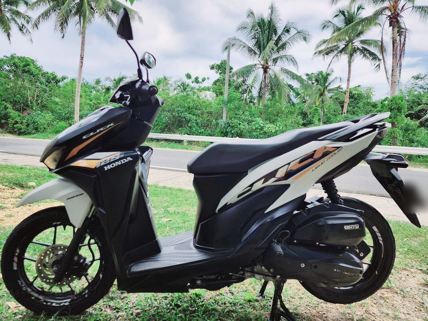 Black and white Honda Click 125i scooter parked on grass beside a palm-lined road