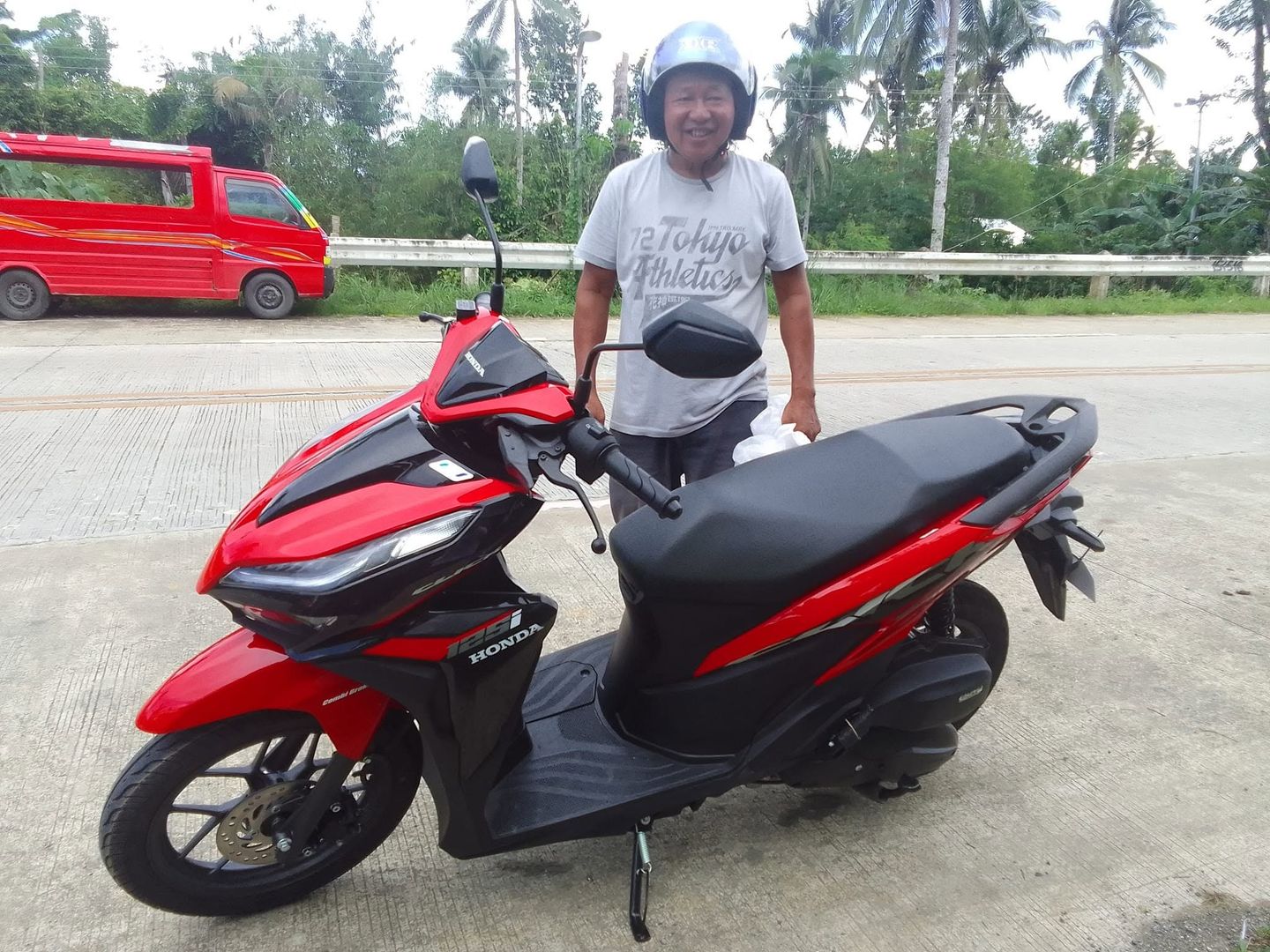 Brand-new red Honda Click 125i with a smiling man in a helmet by the highway