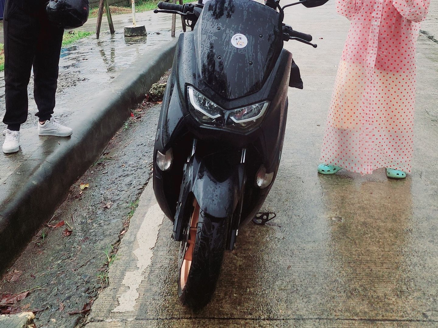 Travellers and a host in a polka-dot raincoat beside a black Yamaha NMAX on a rainy day