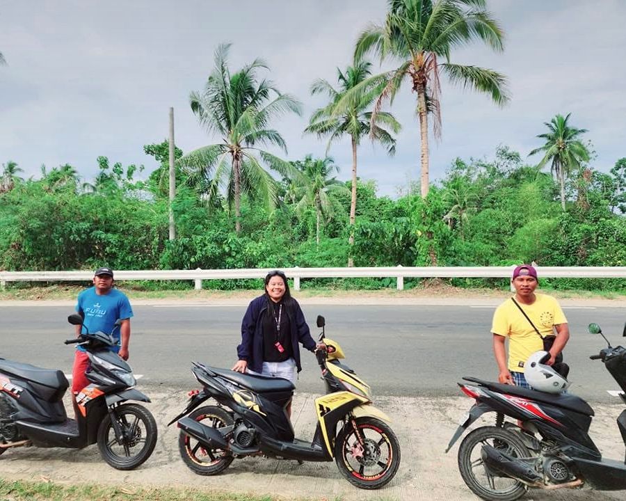 Three members of the Siargao Rose team standing behind rental scooters on a palm-lined highway