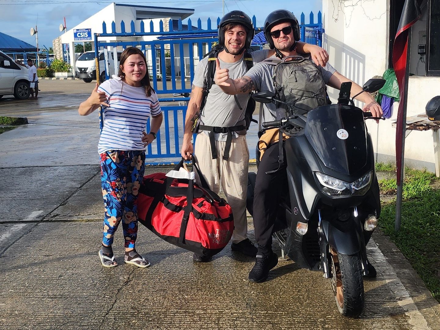 Two travellers in helmets and a smiling woman giving thumbs-up beside a black Yamaha NMAX outside the airport gate